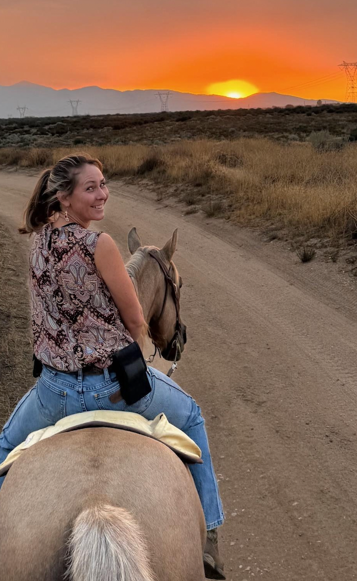 A woman is riding a horse down a dirt road at sunset.