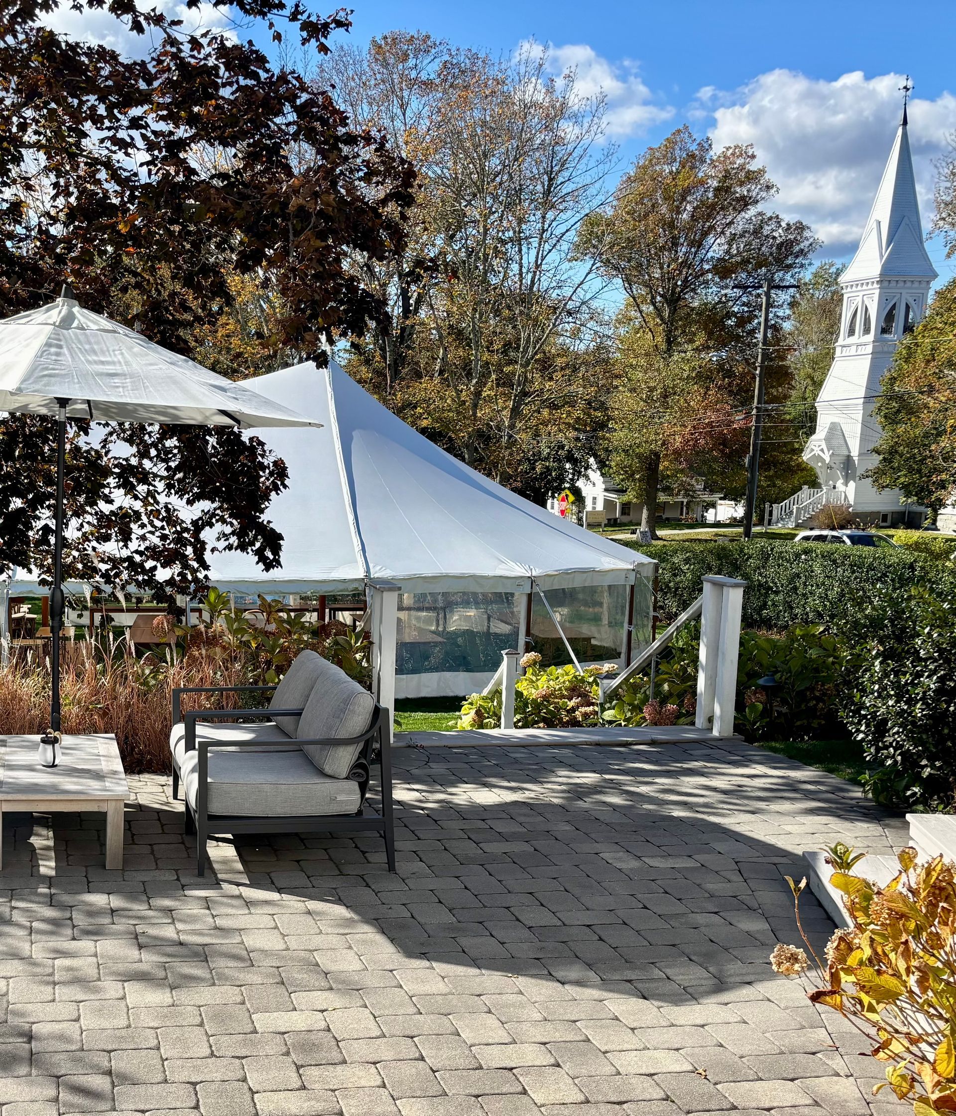 Outdoor patio with tents, seating, trees, and a white church steeple in the background.