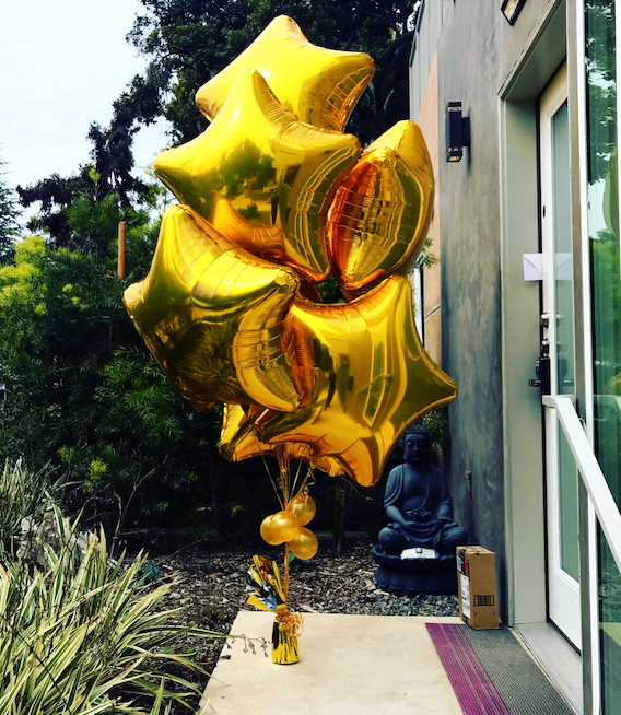 Gold star-shaped balloons tied with a gold ribbon, in front of a building with a Buddha statue.