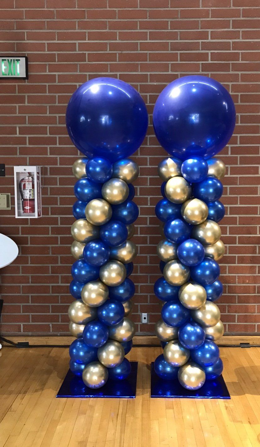 Two balloon columns with gold and blue balloons, topped with large blue balloons, set against a brick wall.