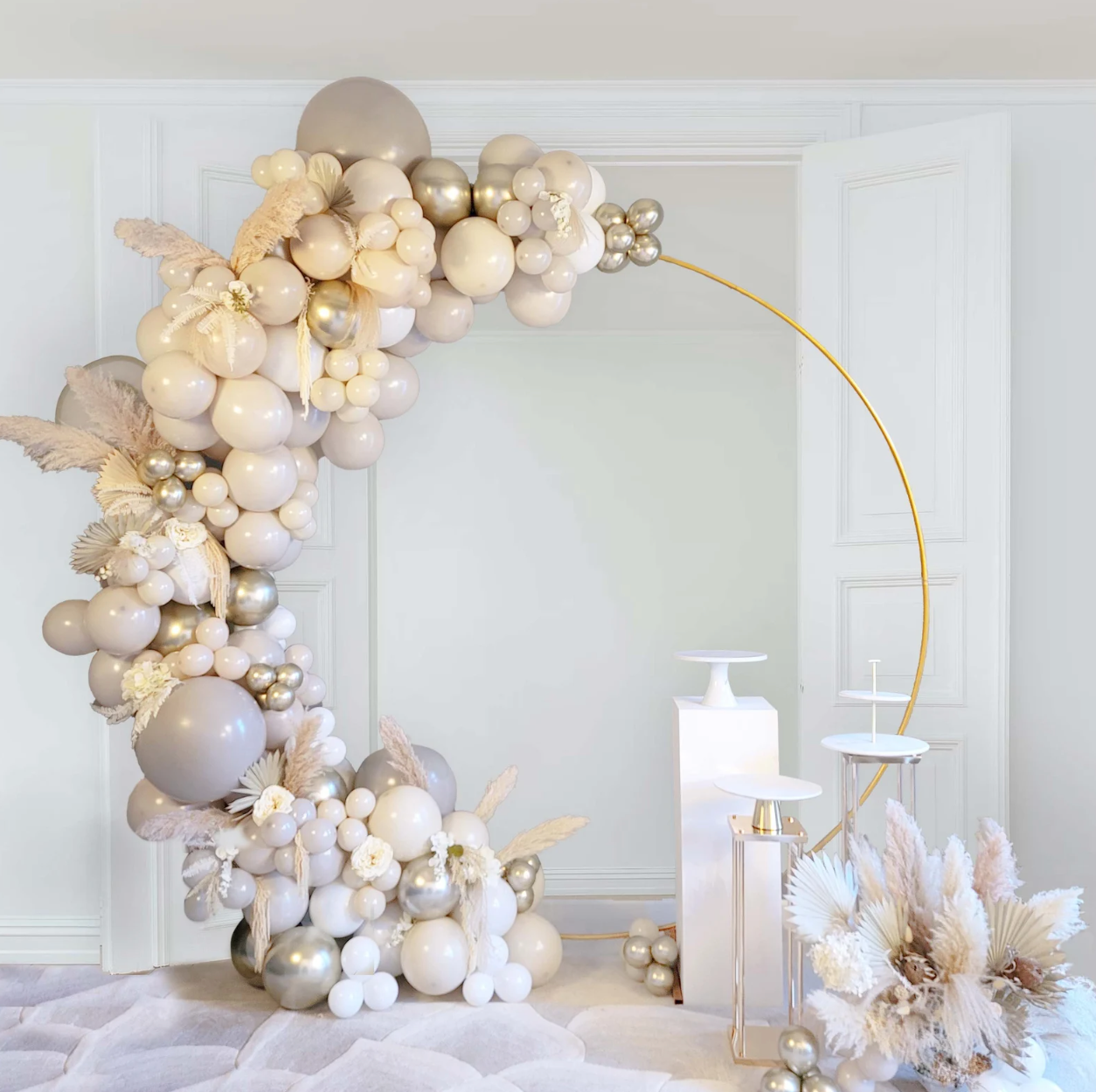Balloon arch with silver, white, and gray balloons. Gold circular frame, white pedestals, and pampas grass.