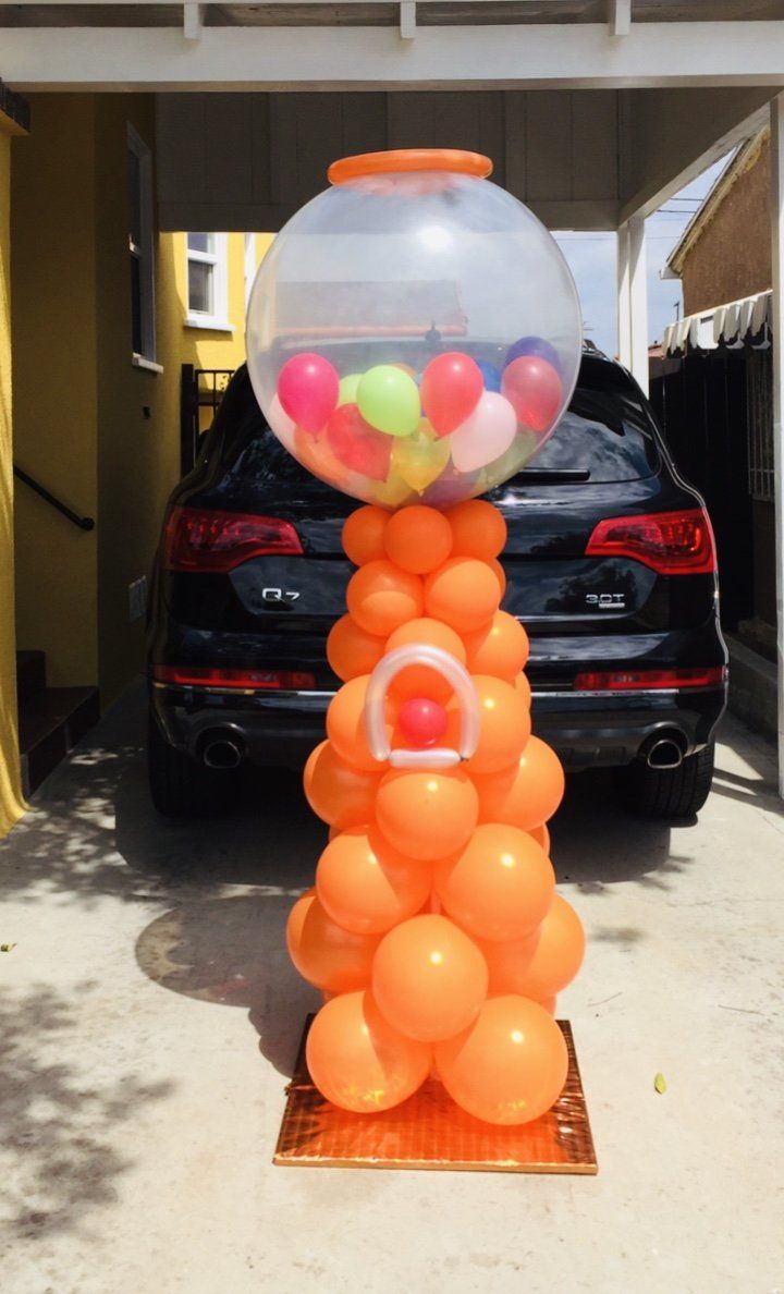 Orange balloon gumball machine with clear top full of colorful balloons, set outdoors.