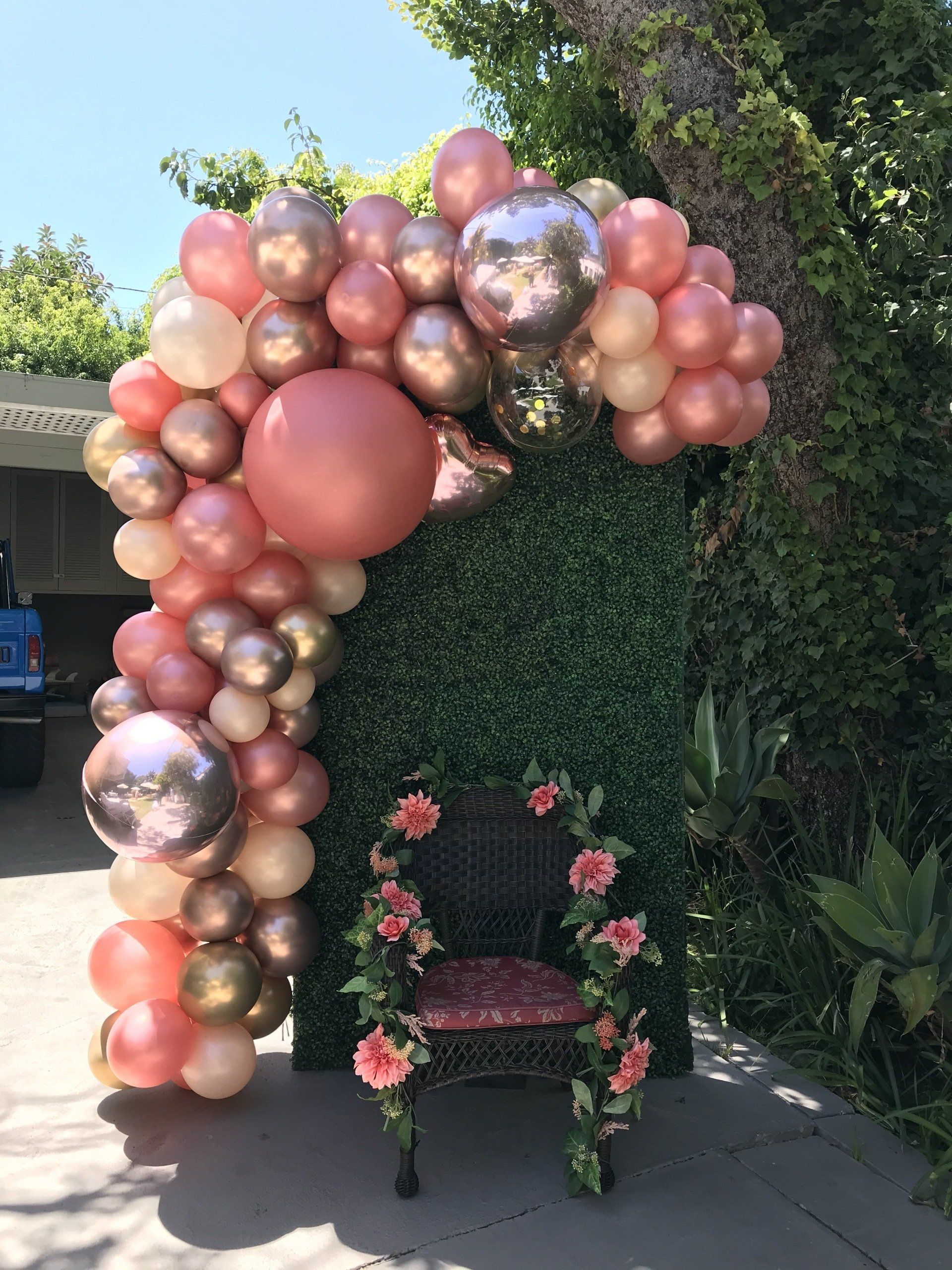 Rose gold and pink balloon arch with green hedge backdrop and chair decorated with flowers.