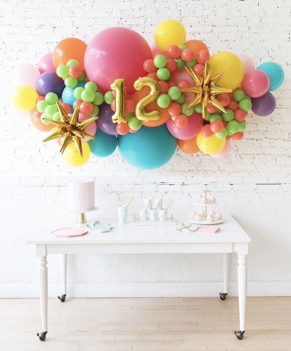 Colorful balloon arch with gold “12” above a white table with party supplies.