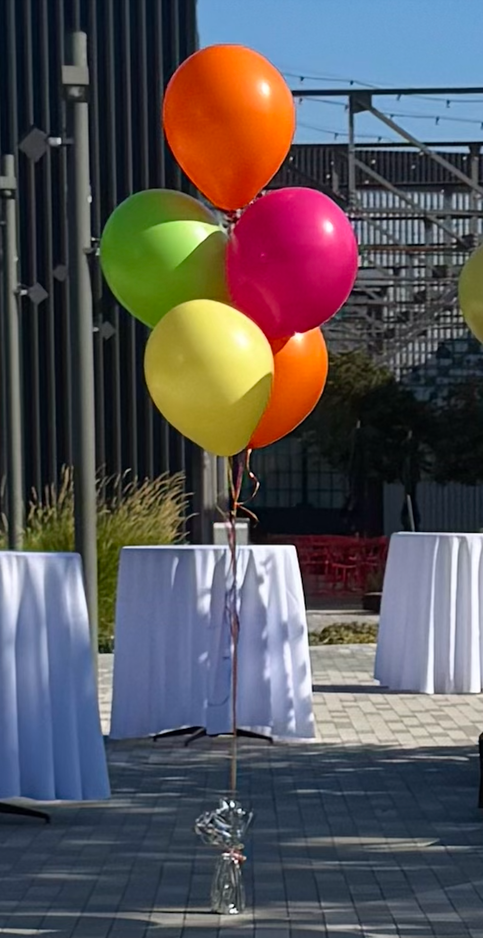 Bunch of colorful balloons tied to a stand above white-covered tables outdoors.
