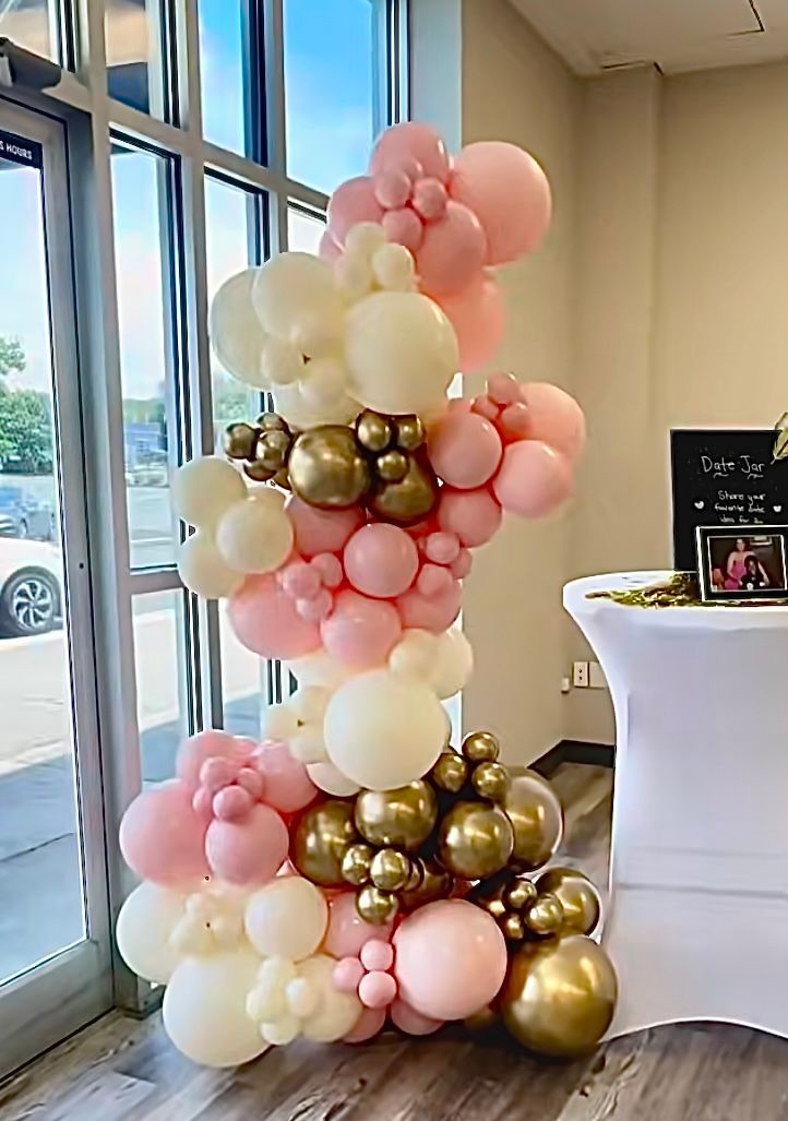 Balloon column in pink, cream, and gold, next to a window and table with a white cloth.