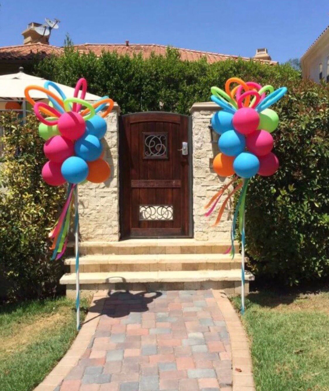 Two colorful balloon clusters flank a wooden door, standing on posts. Bright balloons, ribbon accents, sunny day.