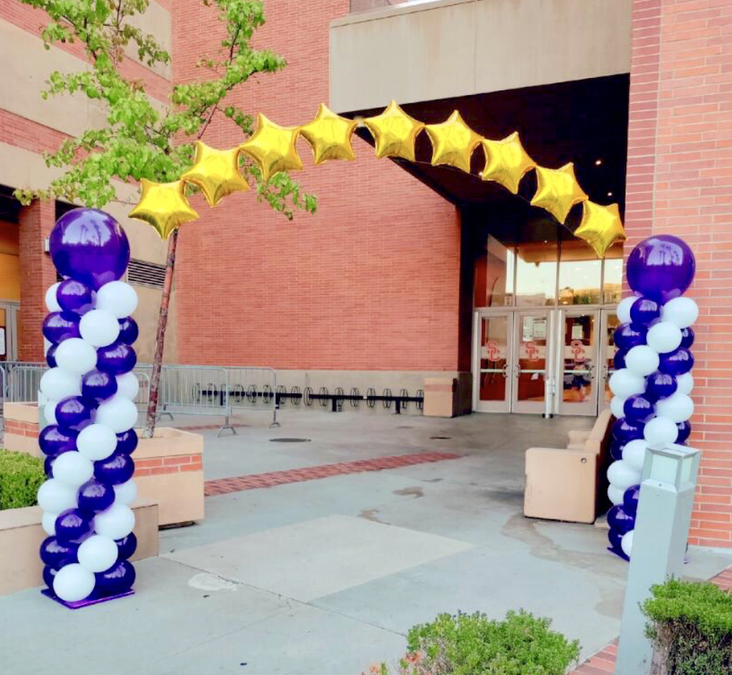 Entrance decorated with purple, white, and gold balloons. Star archway and pillar decorations outside a brick building.