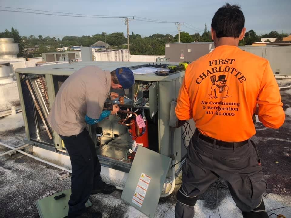 Two men are working on an air conditioner on a roof.