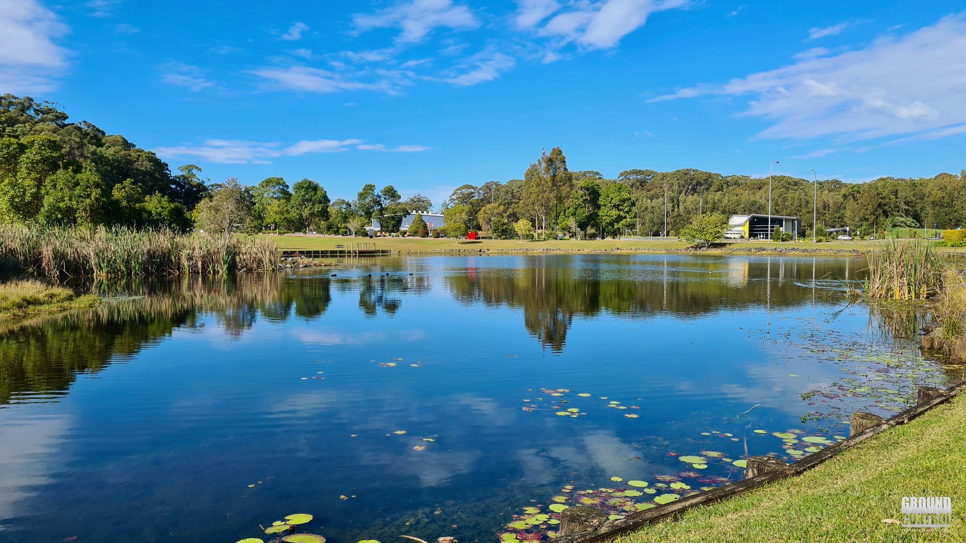 Calm Lake Reflecting Blue Sky, Trees, and Grassy Shoreline on A Sunny Day — Ground Control Property Maintenance In North Boambee Valley, NSW