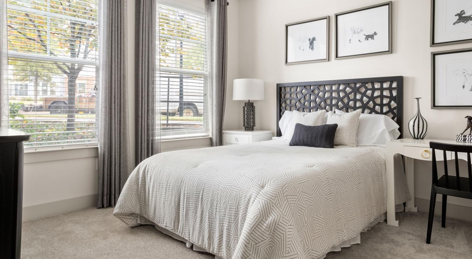 A bedroom with a bed , desk , lamps and two windows.