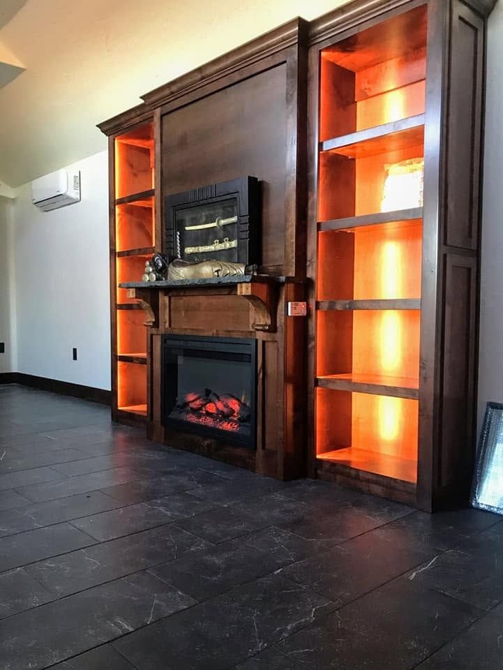 Empty room with a dark wood fireplace, glowing firebox, and orange-lit built-in shelves.