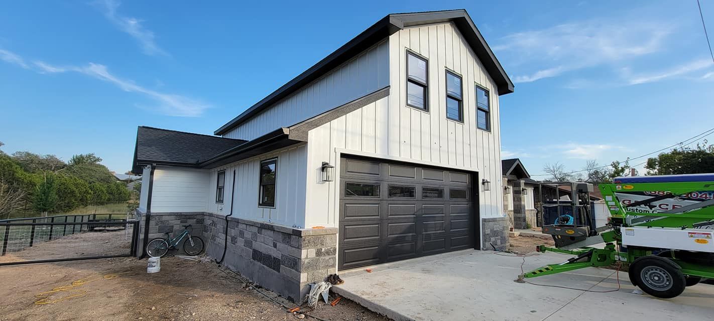 Modern two-story white house with black garage doors and stone base on a dirt lot under a blue sky