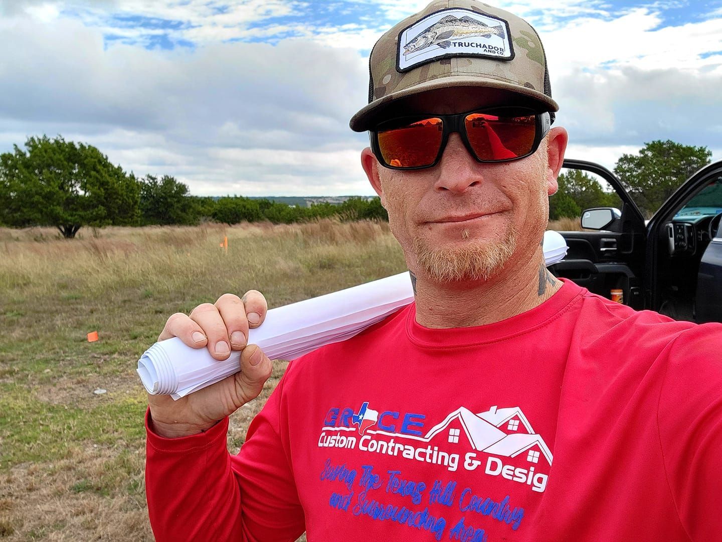 Man in red shirt and cap holds rolled paper outdoors beside an open vehicle