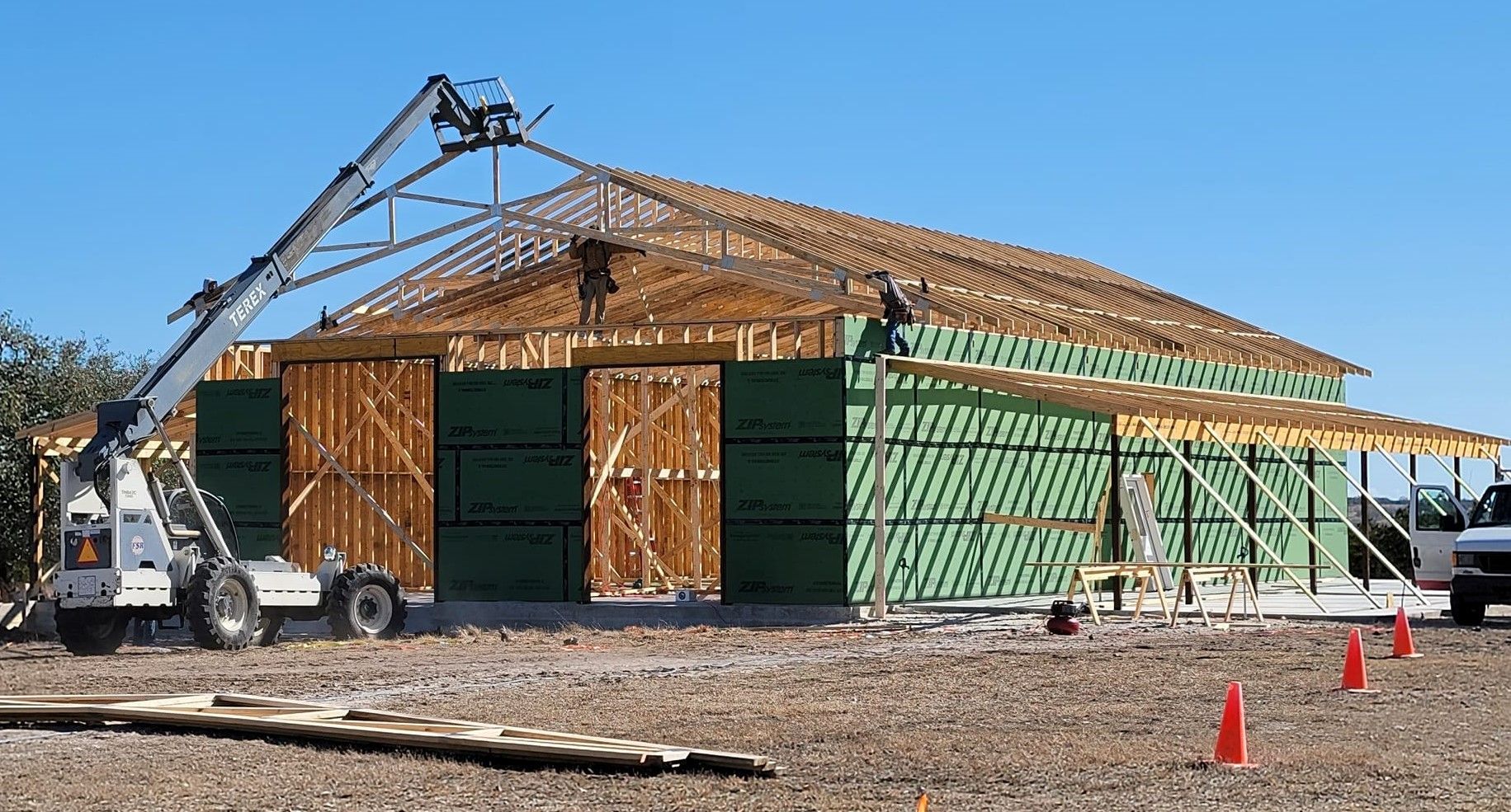 House under construction with wooden roof framing, green sheathing, and a crane on a sunny lot