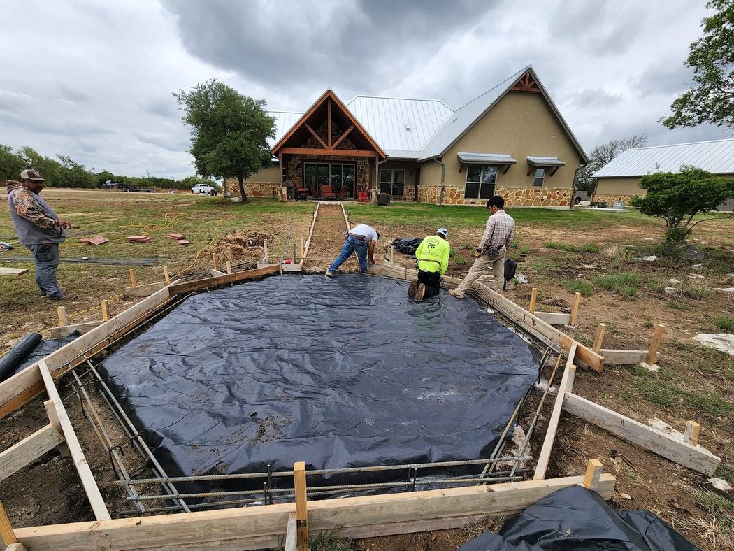 Workers install black pond liner in a dug-out basin at a rural home construction site.