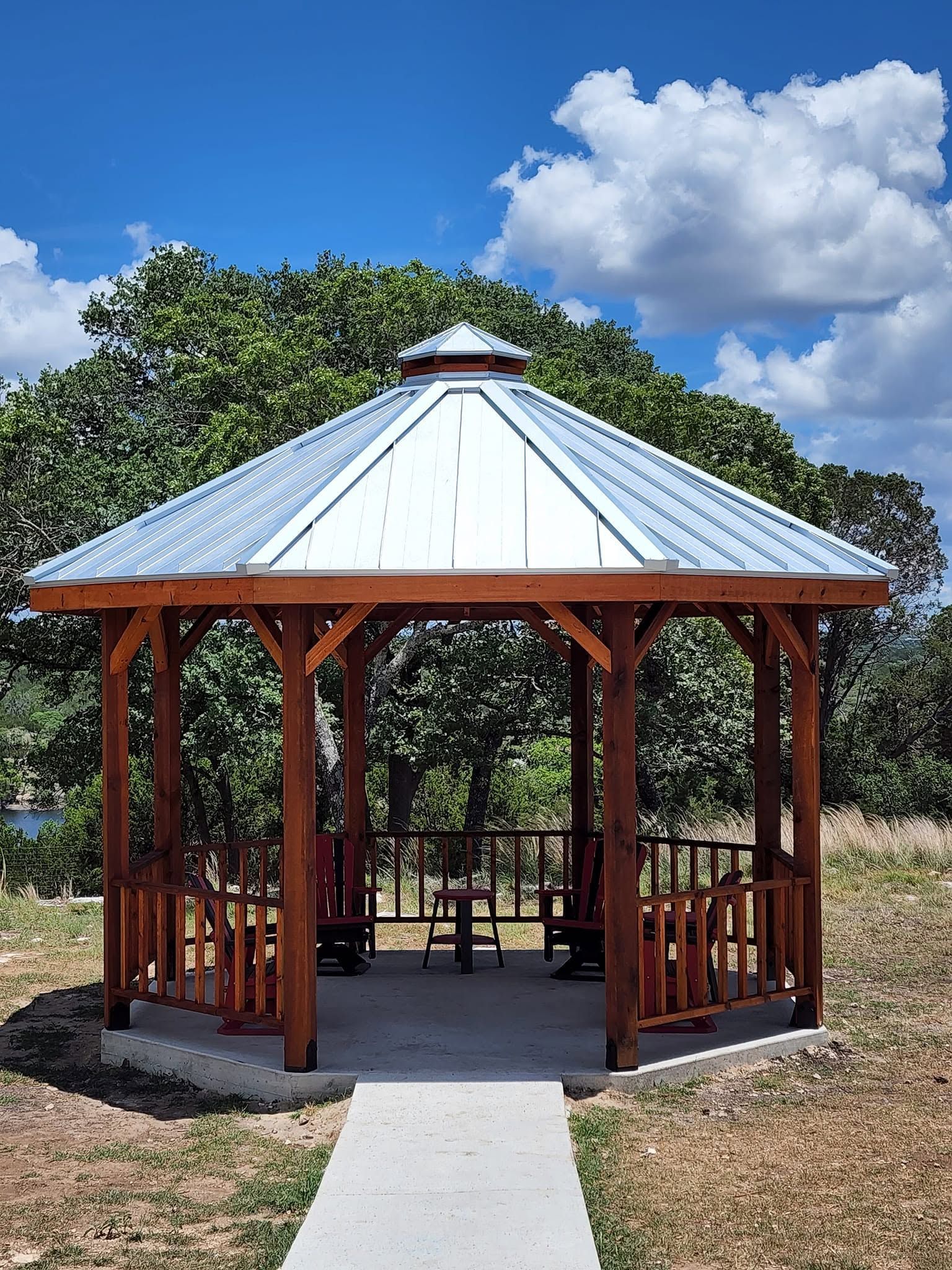 Wooden gazebo with a metal roof and picnic table in a grassy park under a blue sky.