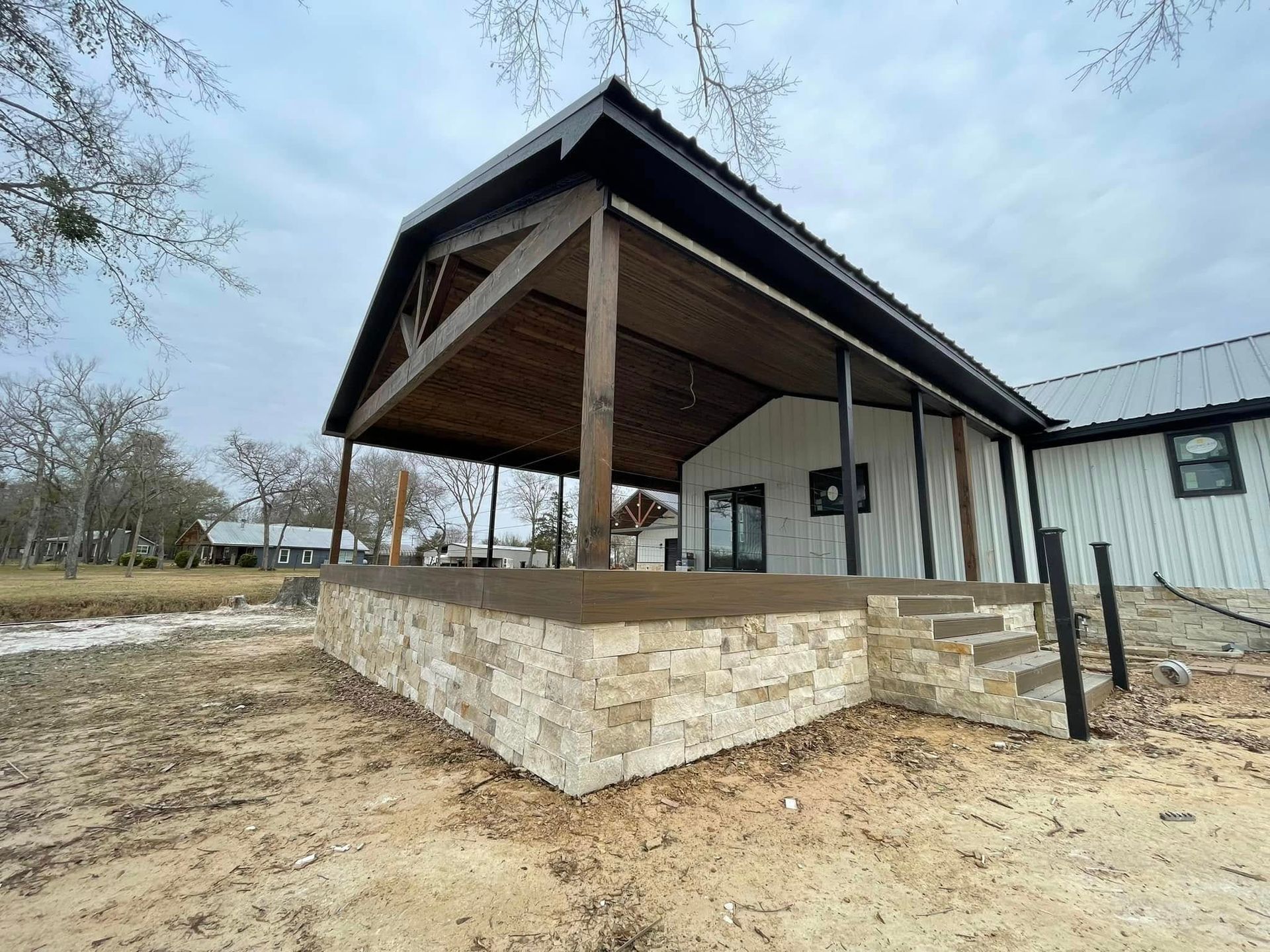 Covered patio with stone retaining wall beside a white building on a dirt lot under a cloudy sky