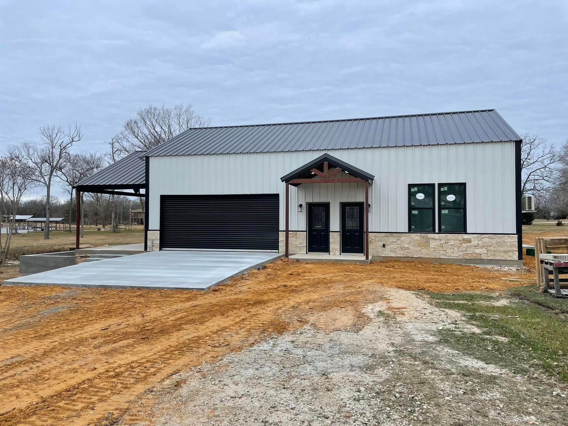 New white metal building with black garage door and covered entry, on a dirt lot under cloudy skies