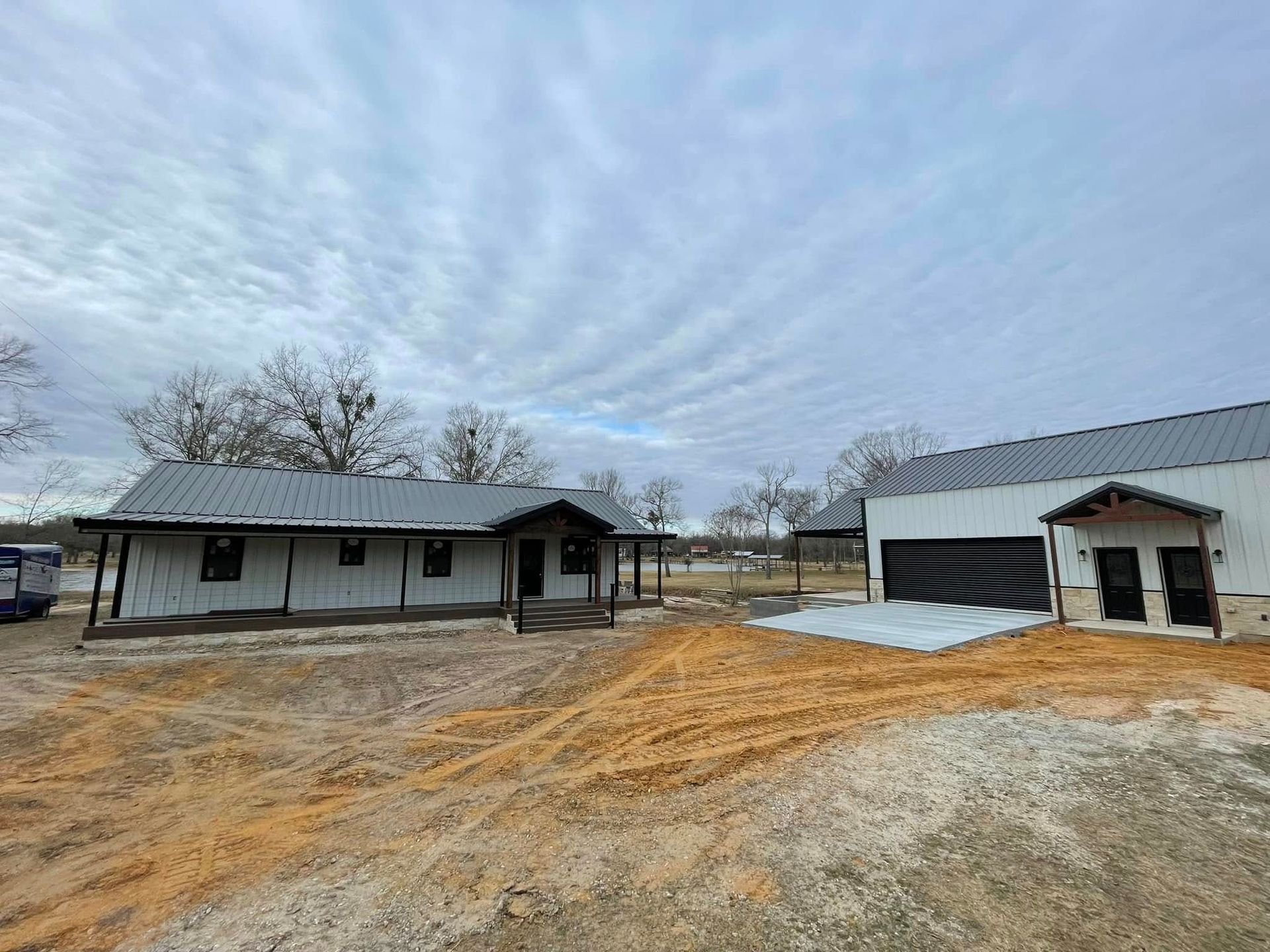 Rural property with a small gray house, large metal shed, and bare yard under a cloudy sky
