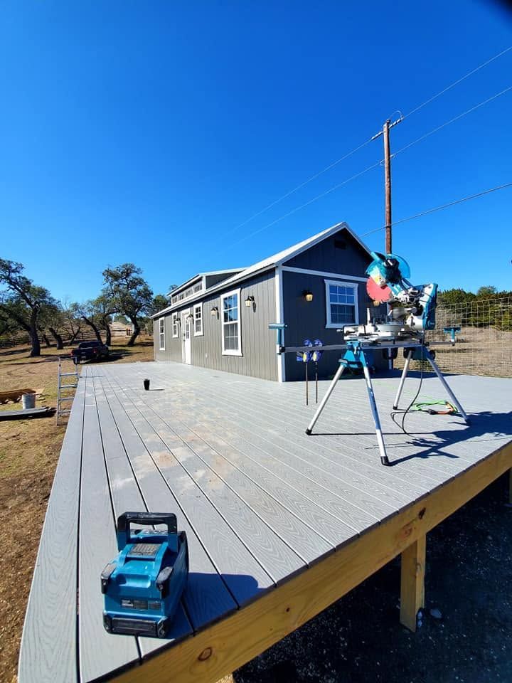 Tiny house on raised deck under clear blue sky, with a ladder, tools, and construction materials nearby.