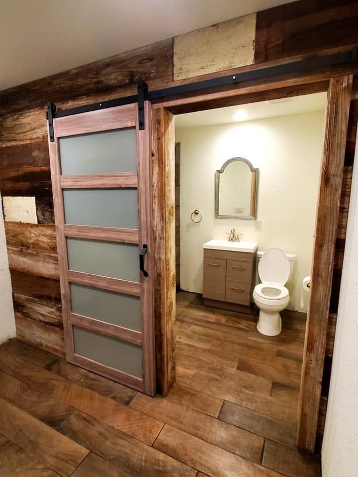 Rustic cabin bathroom with sliding barn door, wood walls and floor, sink, mirror, and toilet.