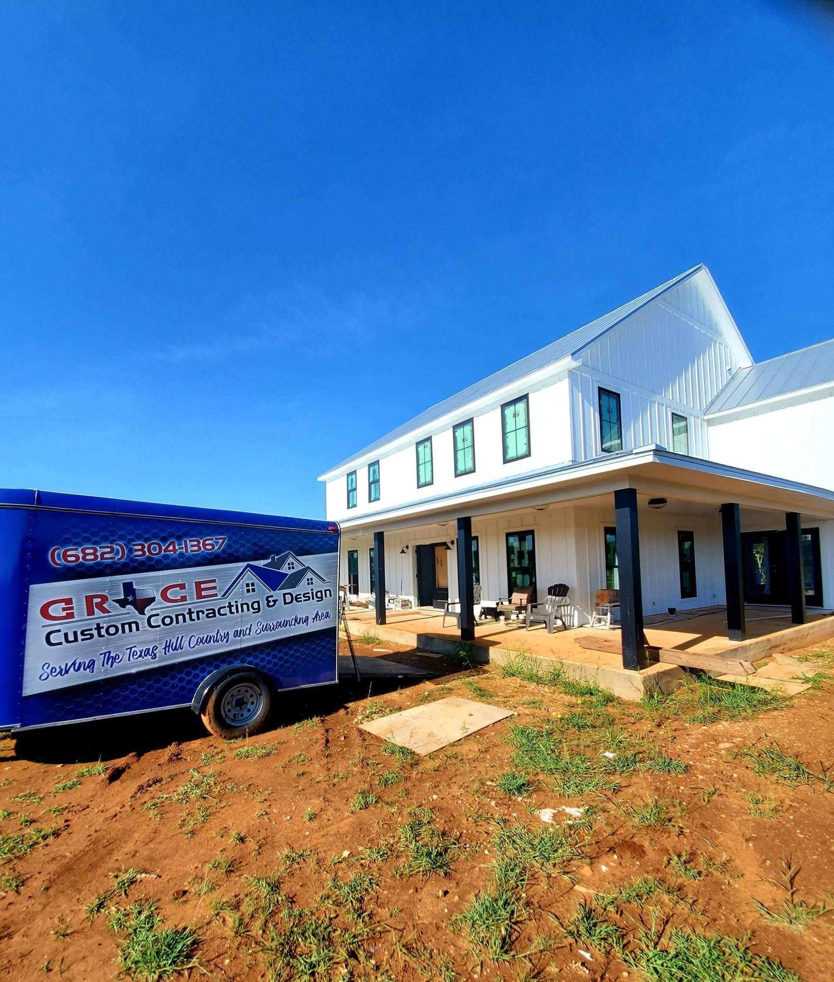 White two-story house under construction beside a blue custom construction trailer on a sunny day