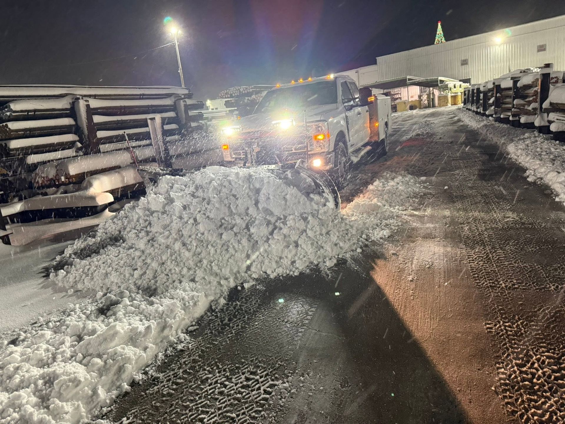 Snowplow clearing a snowy road at night beside parked trucks and a lit building. | 814 Truck & Equipment Services LLC