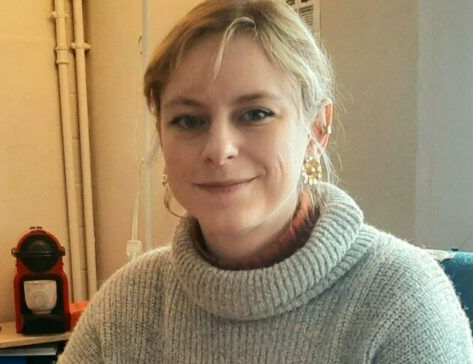 Femme blonde au léger sourire, vêtue d'un pull gris, à l'intérieur. La machine à café est à gauche.