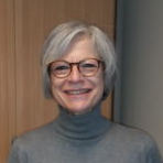 Une femme aux cheveux gris et aux lunettes sourit, vêtue d'un pull à col roulé gris, près d'une porte en bois.