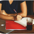 Une personne écrit dans un cahier sur une table en bois avec un ordinateur portable, un classeur rouge et une boisson.
