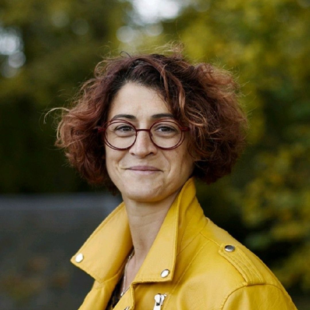 Femme aux cheveux roux bouclés, lunettes, souriante, portant une veste jaune à l'extérieur.