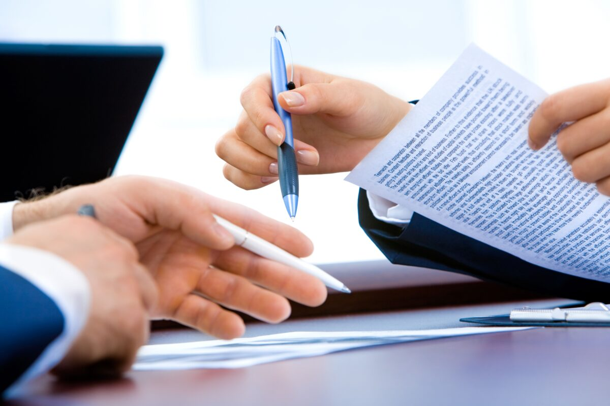 Deux personnes en costume examinent des documents, l'une offrant un stylo à l'autre pour qu'elle les signe.