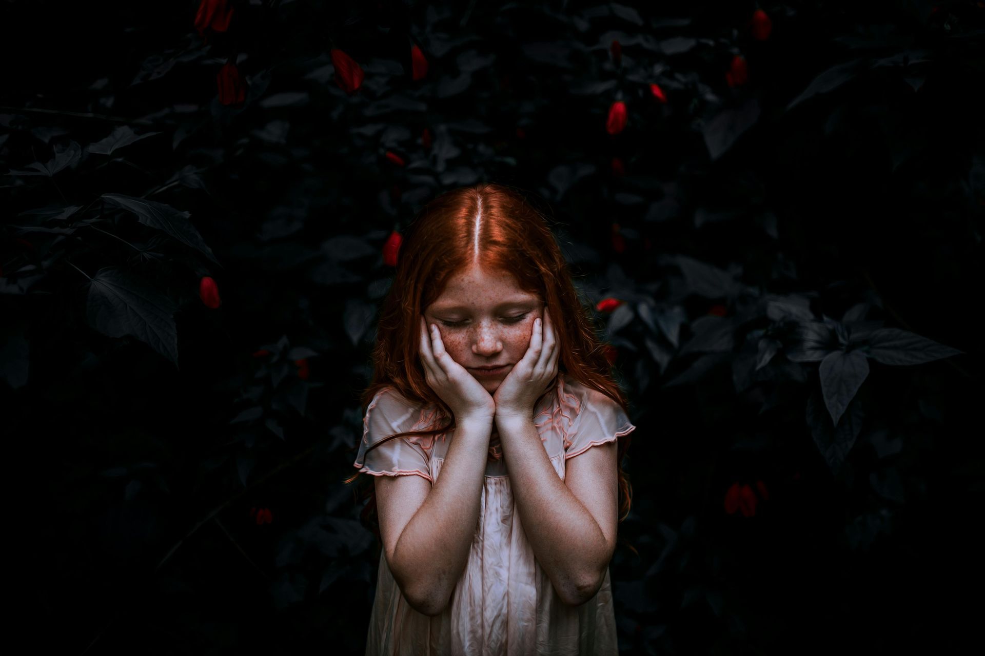 Fille aux cheveux roux, les mains sur le visage, apparaît triste, sur fond de feuillage sombre.