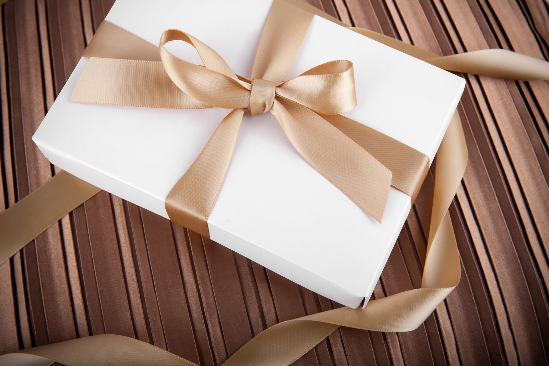 Scatola regalo bianca con nastro dorato su superficie marrone a strisce.