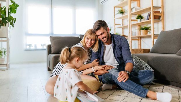 Family of three sitting on a rug in a living room, looking at as book together.