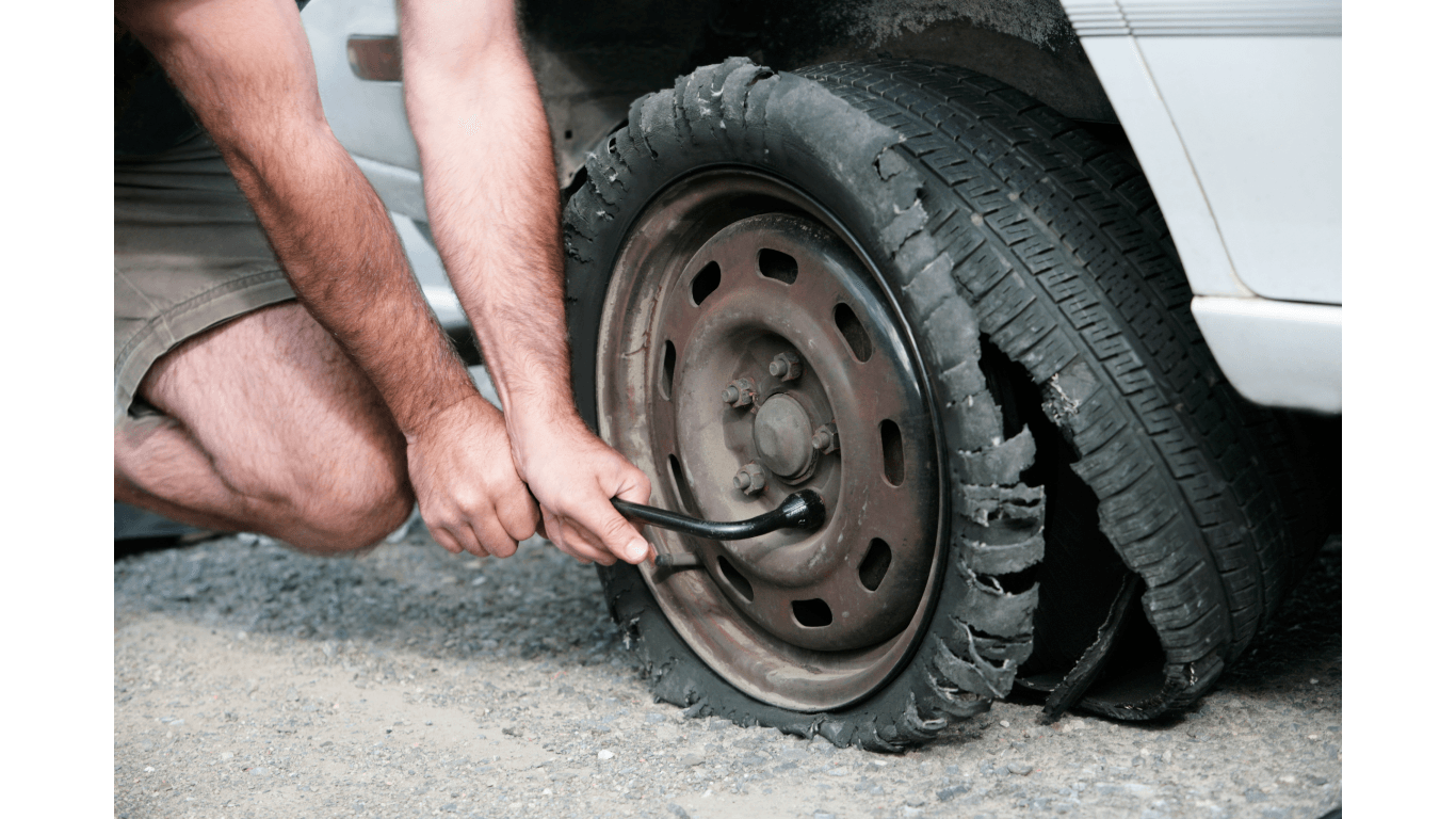 A person uses a lug wrench to loosen the nuts on a car's flat, damaged tire while kneeling on the side of the road.