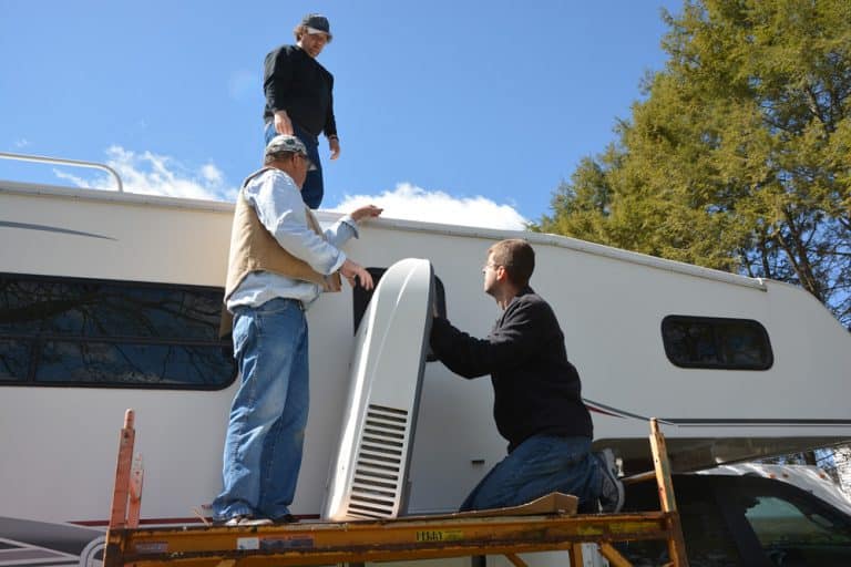 Three people on a scaffold work together to install or remove a large white air conditioning unit on a camper van roof.