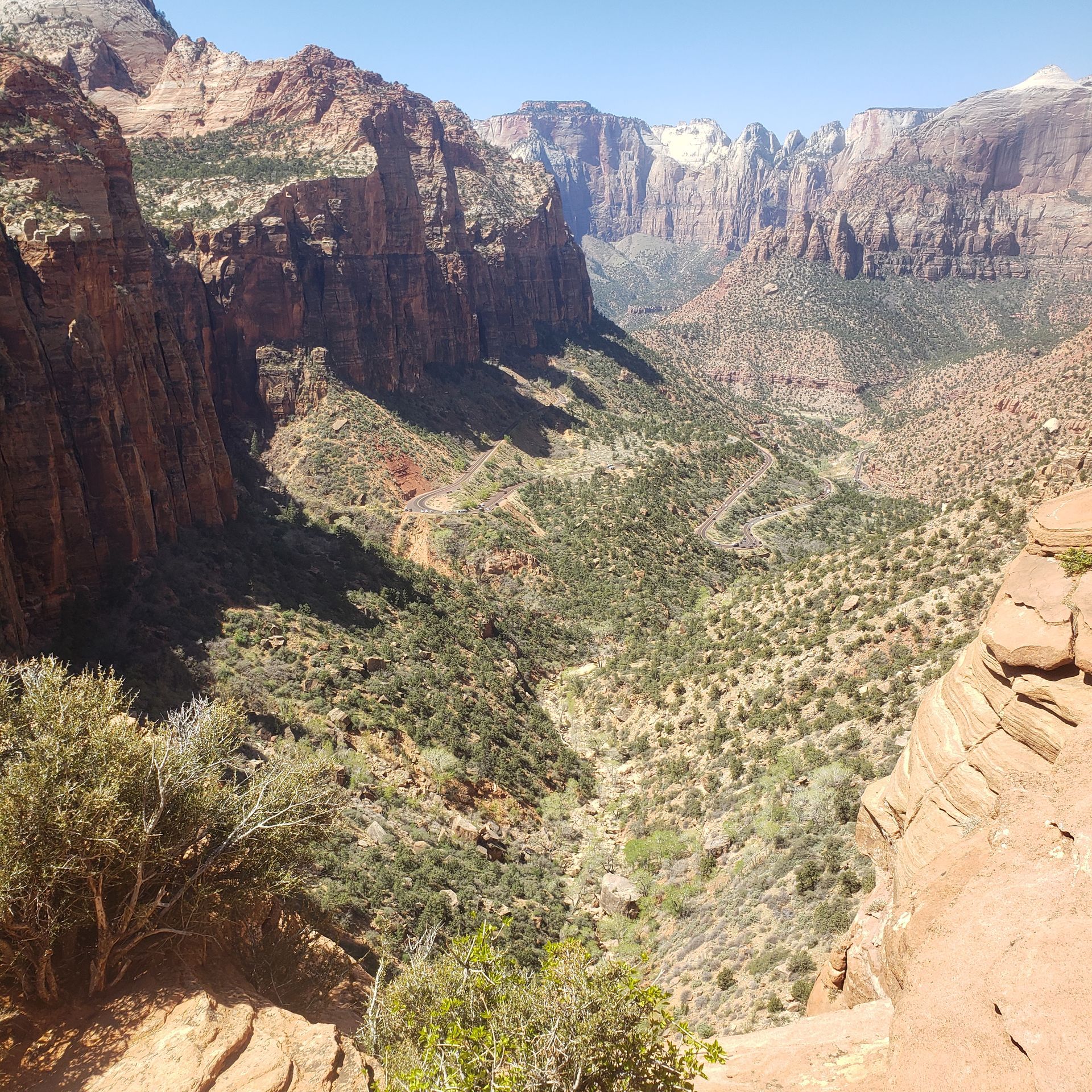 A high-angle view looking down into a deep, rugged canyon with towering red rock cliffs, steep slopes, and desert brush.