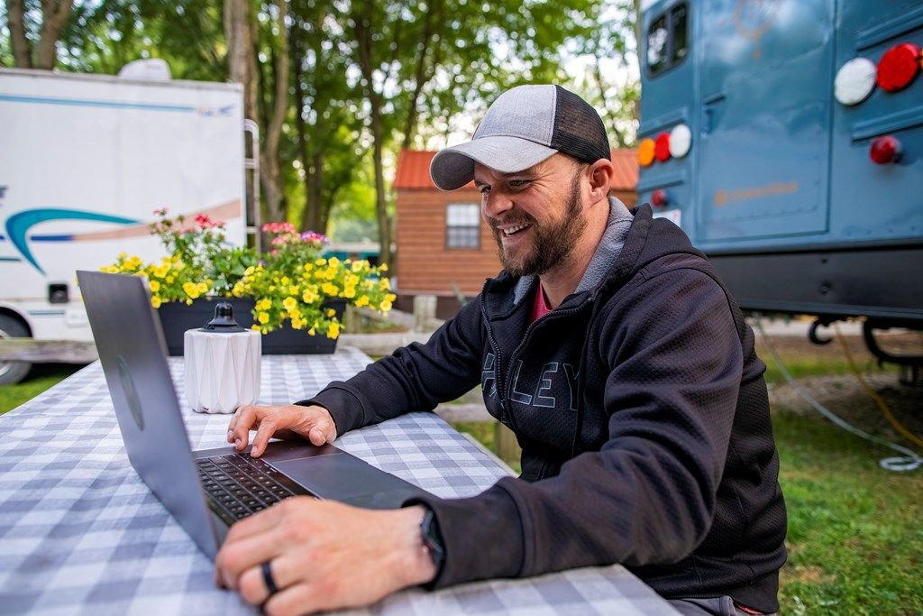 A man in a baseball cap smiles while using a laptop at a table outside near a parked bus and camper.