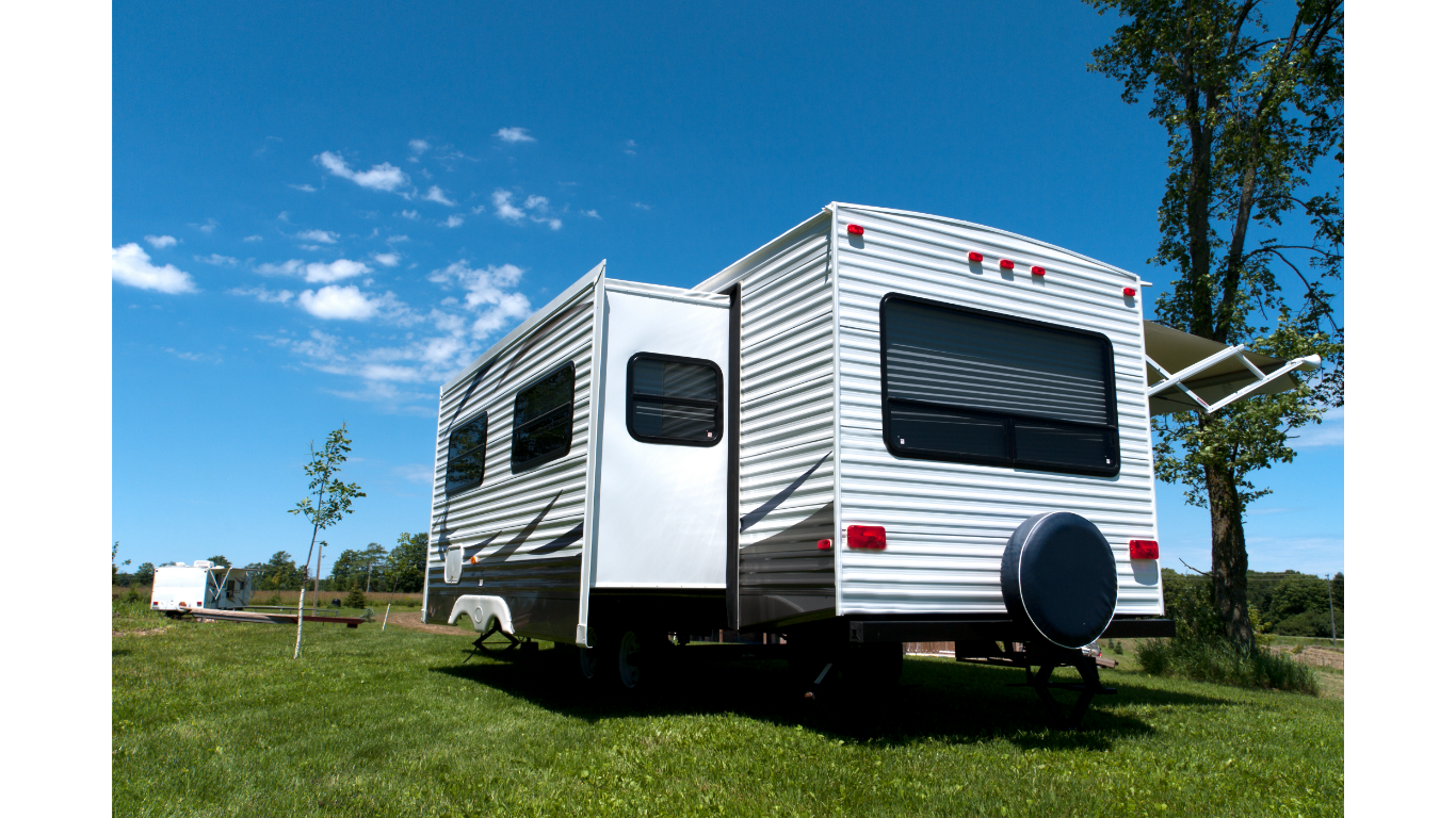 A white travel trailer with a slide-out parked in a grassy field under a bright blue sky.