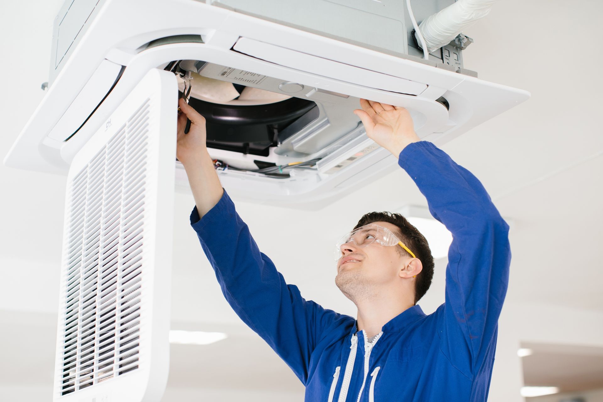 A technician in a blue uniform and safety glasses repairs a ceiling-mounted air conditioning unit.