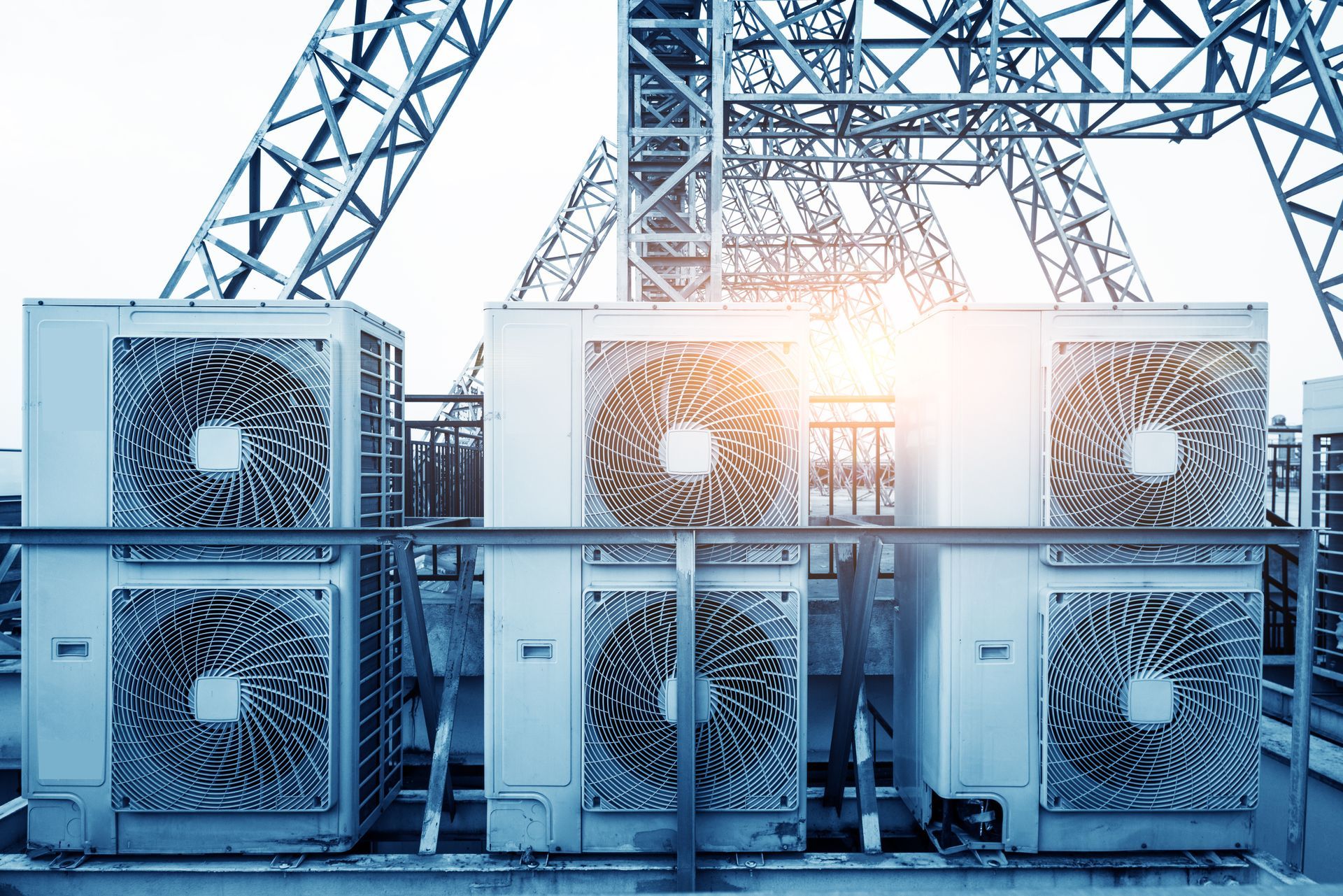 Three industrial HVAC outdoor units arranged on a rooftop beneath a metal scaffolding structure under bright sunlight.