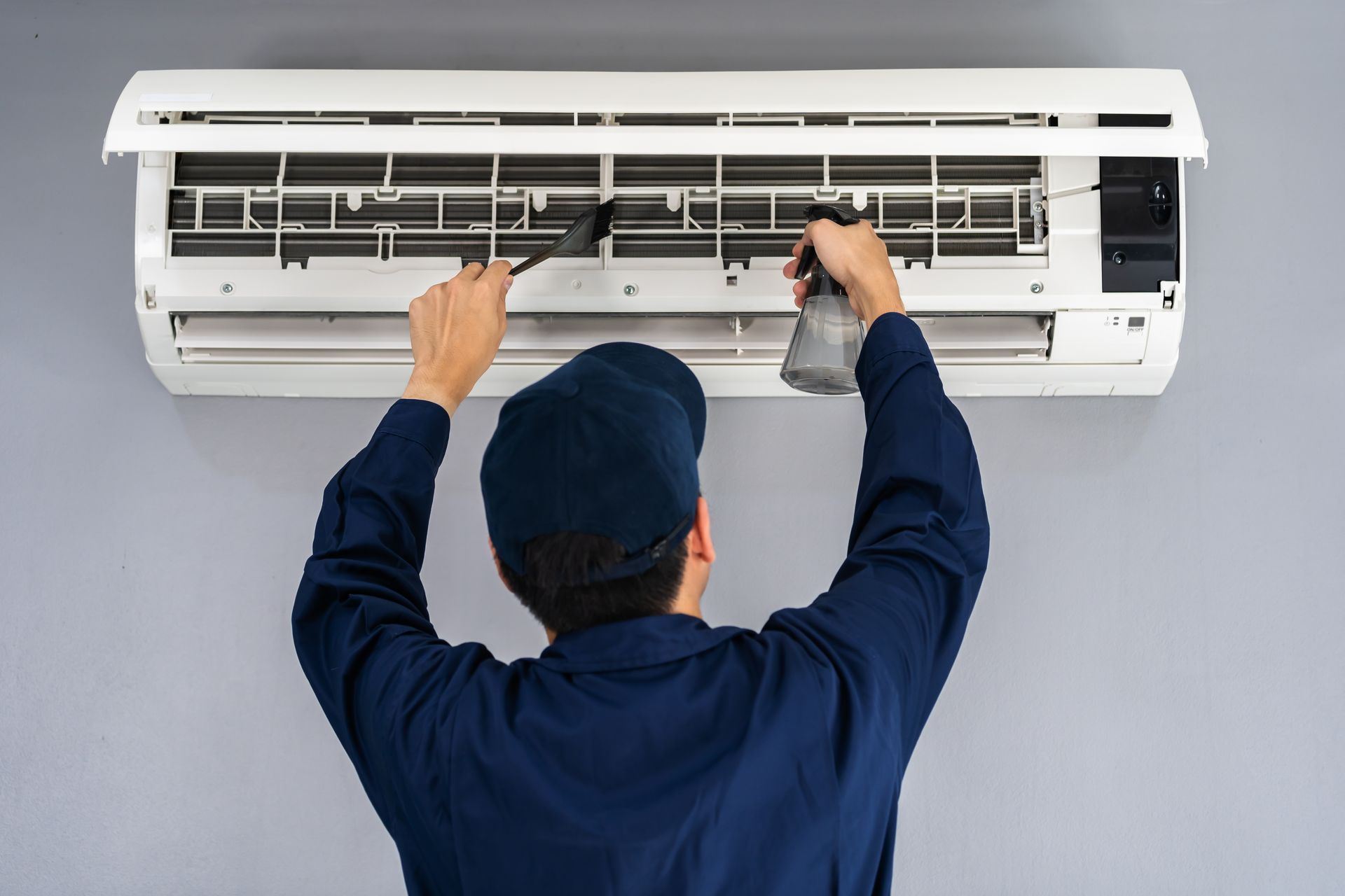 A service technician in a blue uniform cleans the internal components of a wall-mounted air conditioner unit.