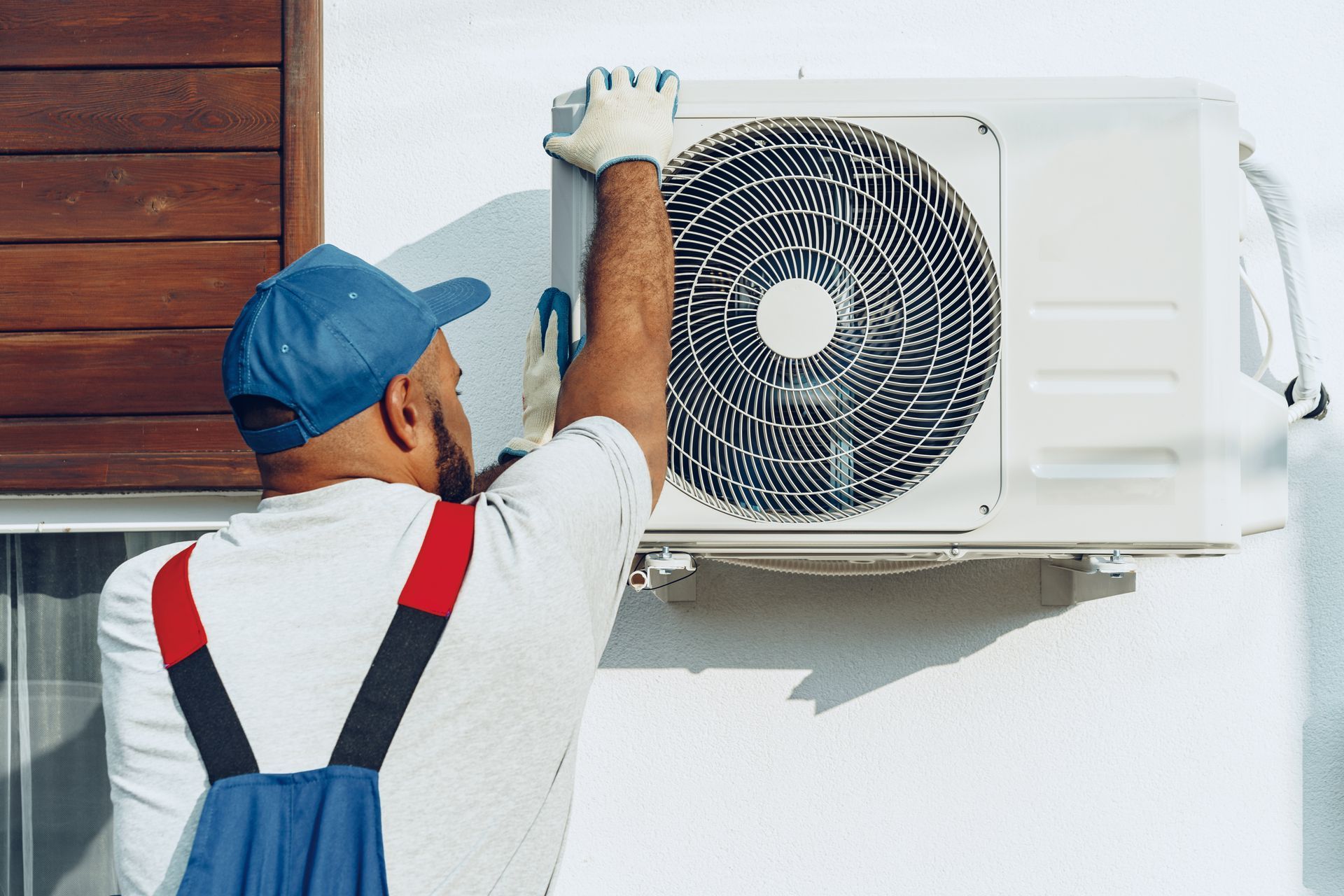A technician in a blue cap and work overalls repairing an outdoor air conditioning unit mounted on a white wall.
