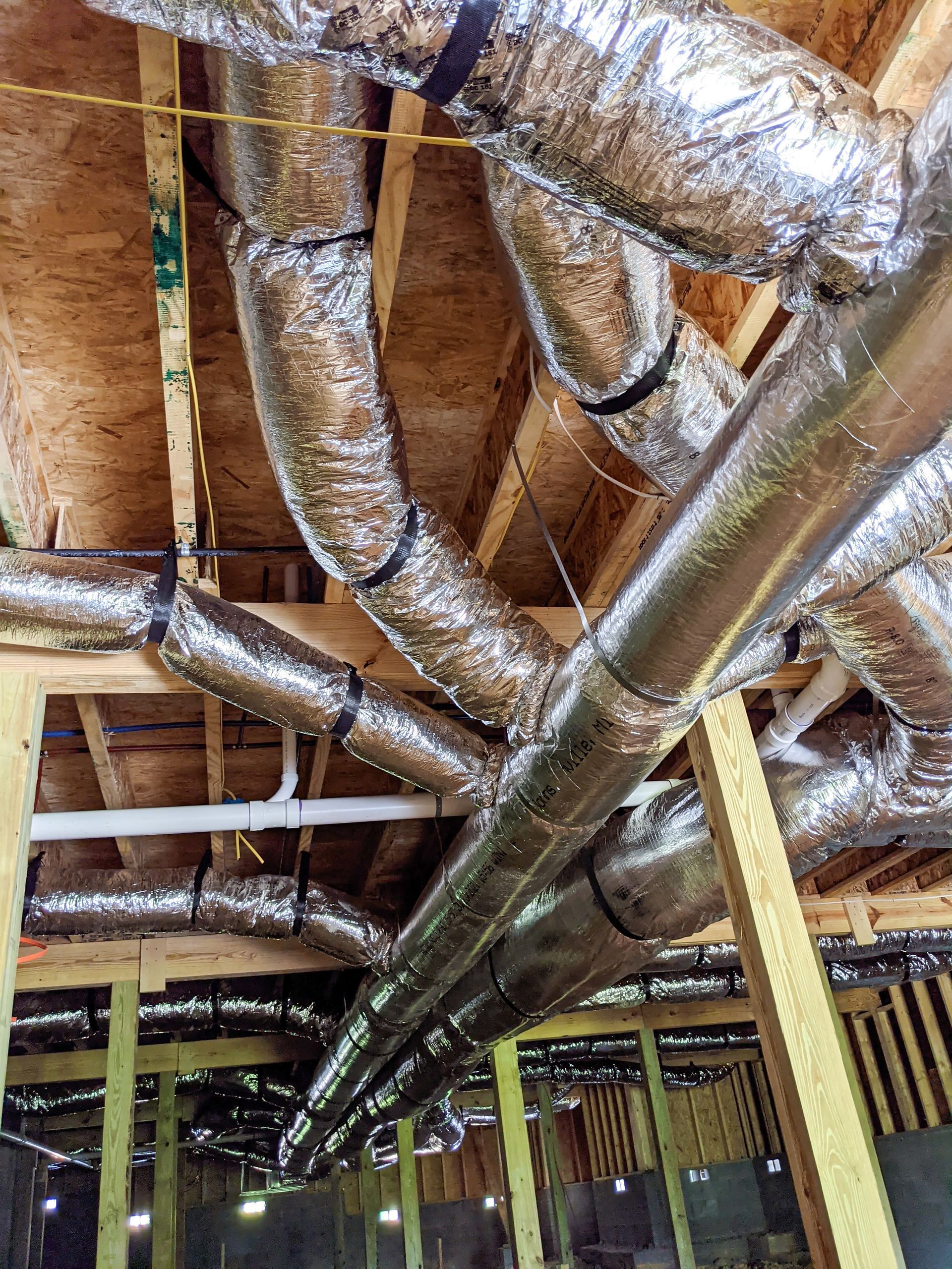 A worker in an attic wearing grey gloves secures silver, foil-insulated ductwork with silver tape.
