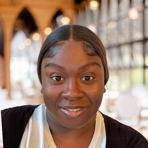 A person with dark hair styled with laid edges smiling at the camera, set against a blurred indoor background with lights.