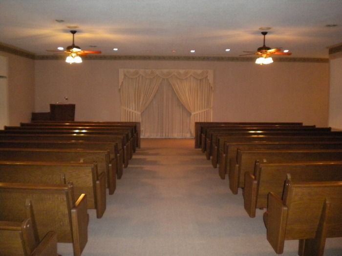 An empty chapel with two rows of wooden pews facing a wall with draped, beige curtains and two ceiling fans.