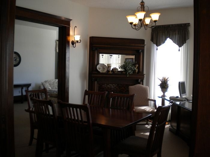 A dining room with a wooden table and chairs, a fireplace with a mirror above it, a chandelier, and an adjacent room.