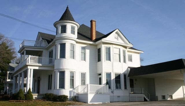 A two-story white Victorian house with a corner turret, wraparound porch, and an attached side carport.